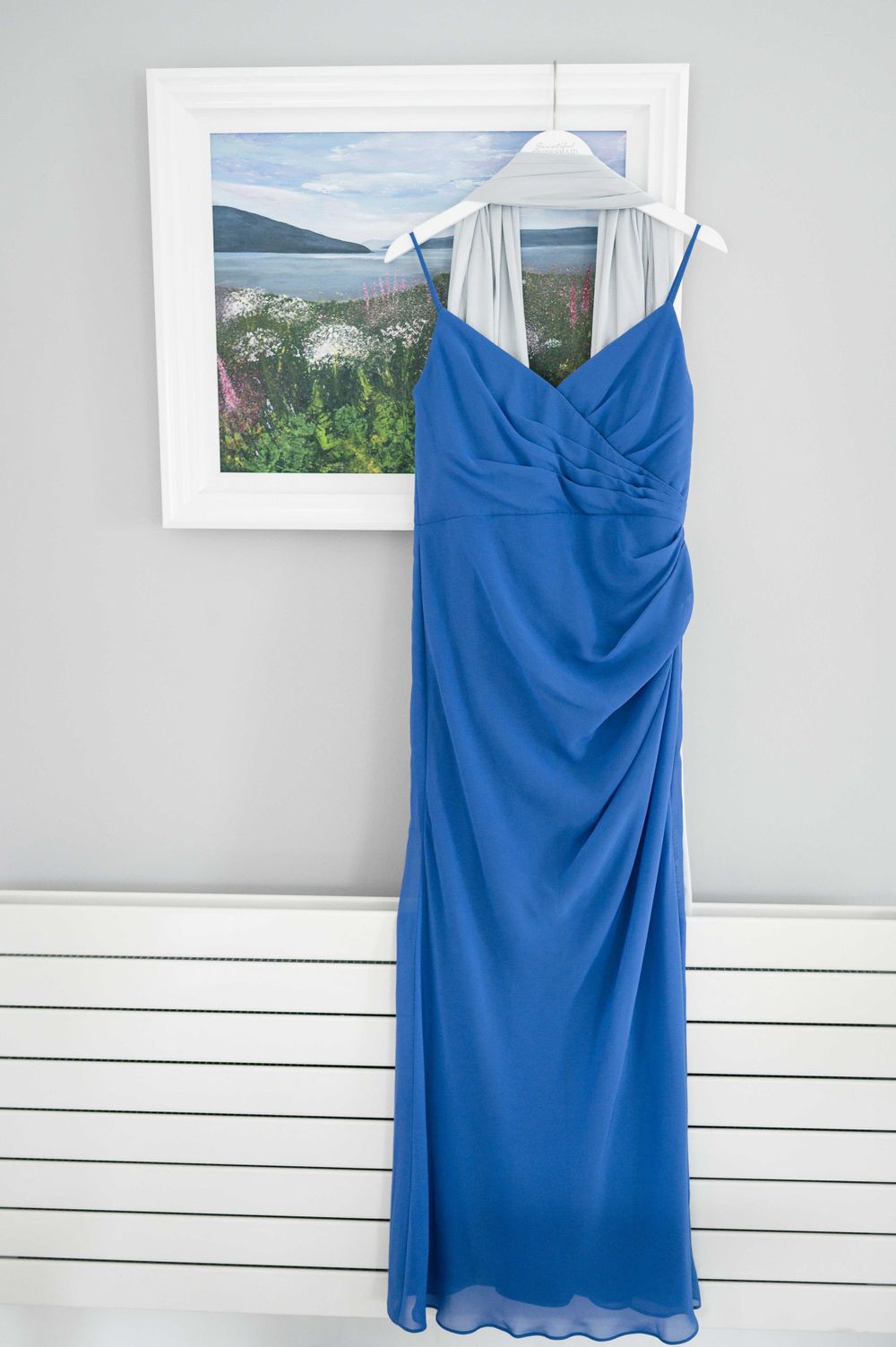 Elegant floor-length blue formal dress hanging on white wall with framed landscape artwork in background.