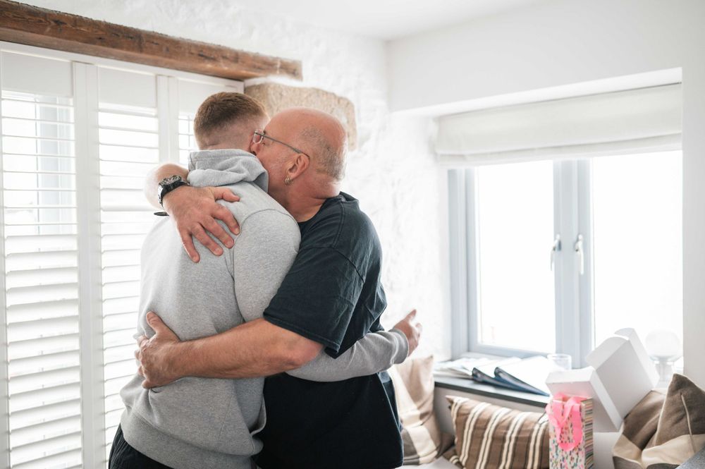 Two people share a warm embrace in a bright living room with white shutters and window light.