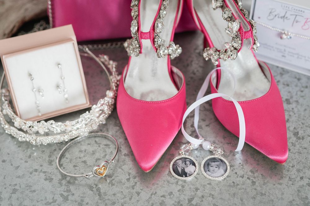 Pink bridal heels displayed with wedding rings, jewelry, and a pearl headpiece on a stone surface.