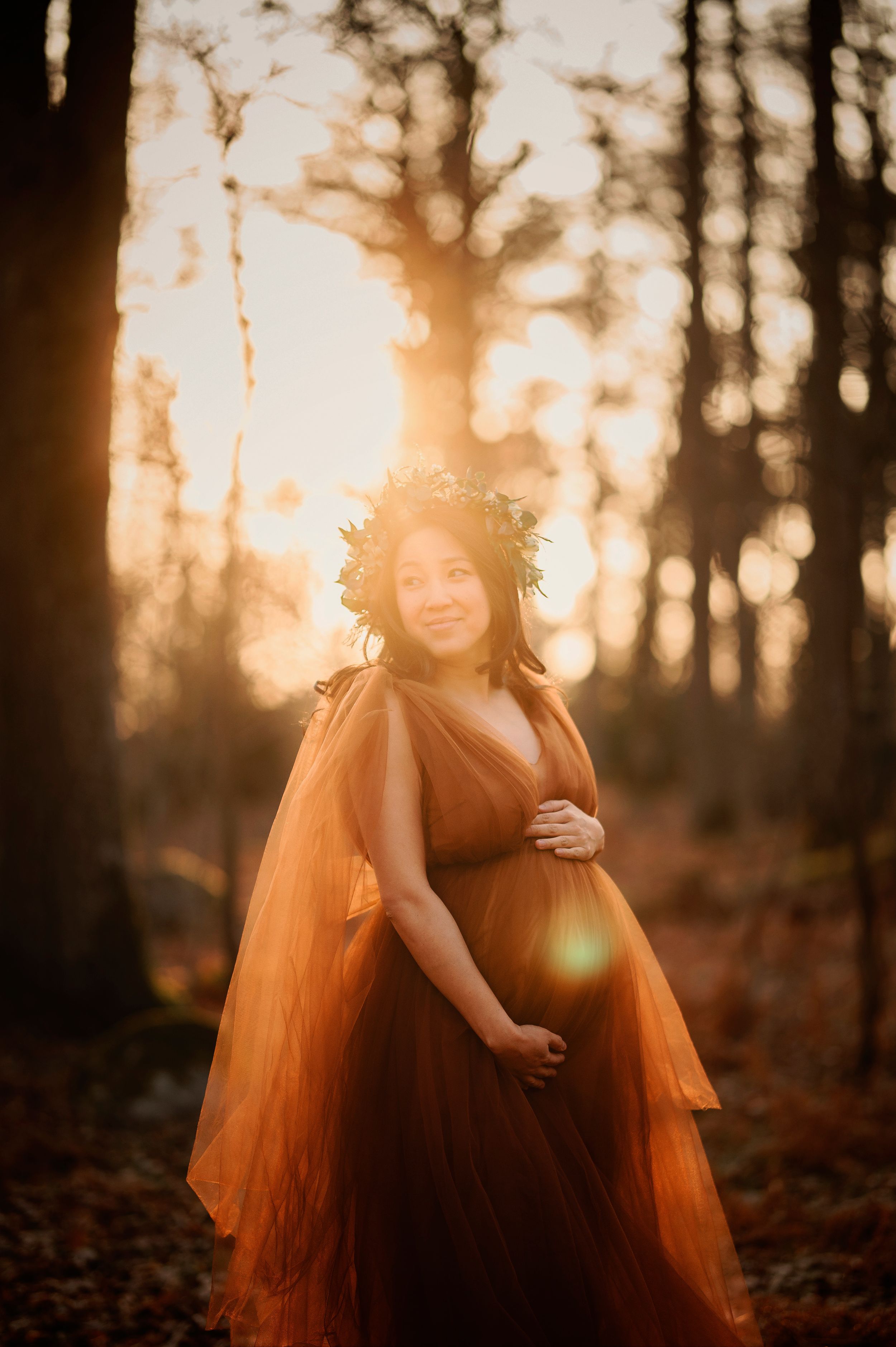 Silhouetted figure in flowy burgundy dress and flower crown poses in sunlit autumn forest during golden hour.