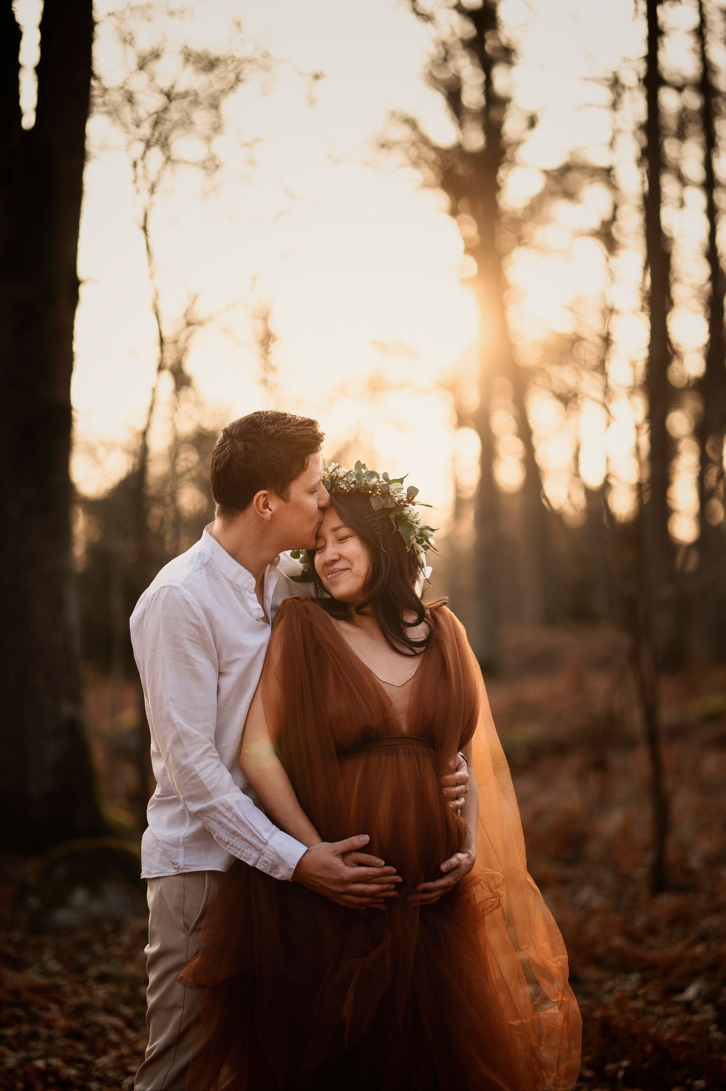 Romantic maternity photo of couple embracing in golden sunlit forest during sunset.