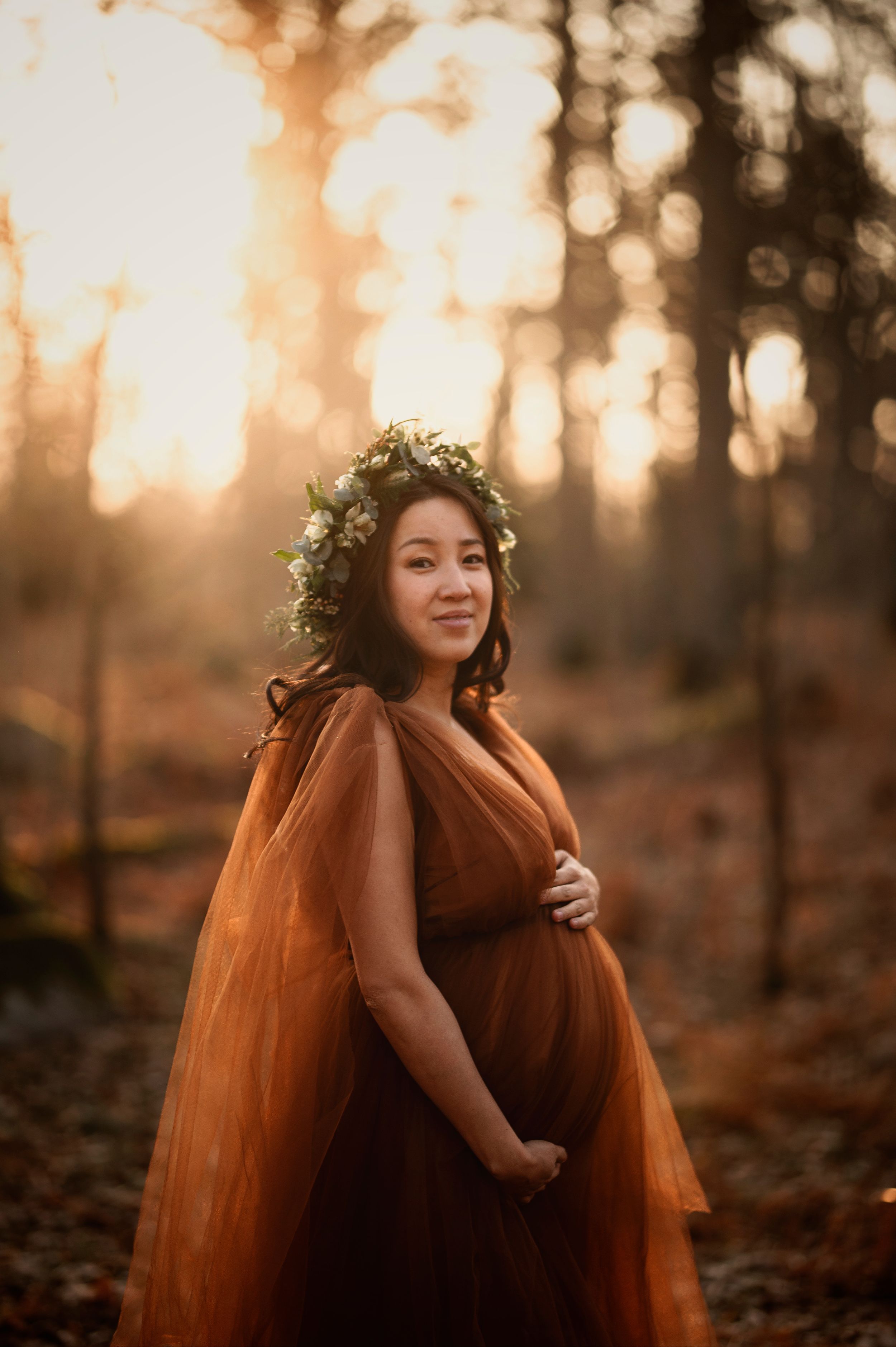 Expectant mother wearing brown flowy dress and floral crown poses in woodland setting during golden hour sunset.