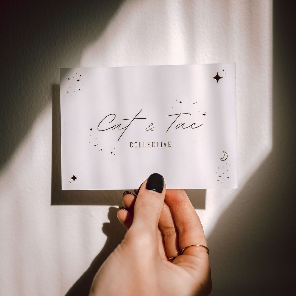 Hand holding minimalist business card with 'Cafe & Tae' text against white wall with shadow pattern.