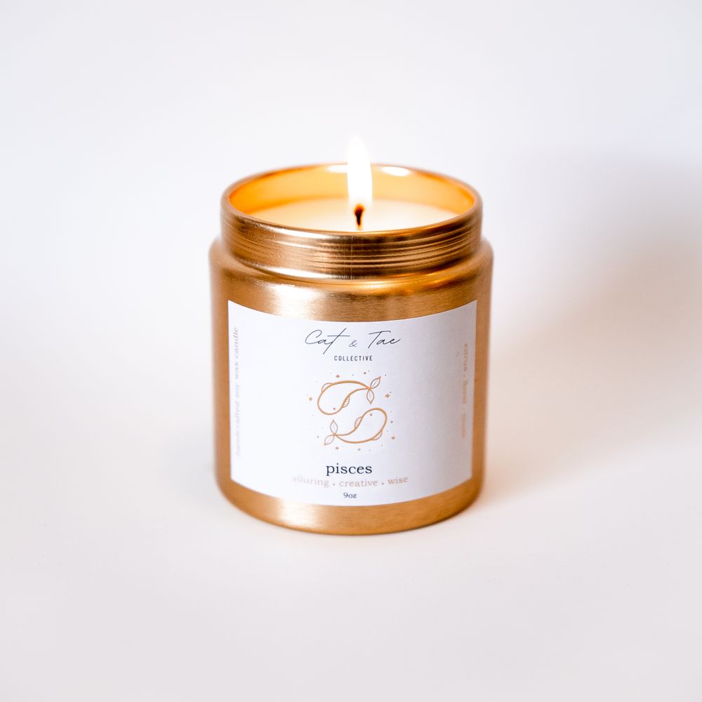 Lit gold zodiac-themed candle in glass jar with minimalist white label against light background.