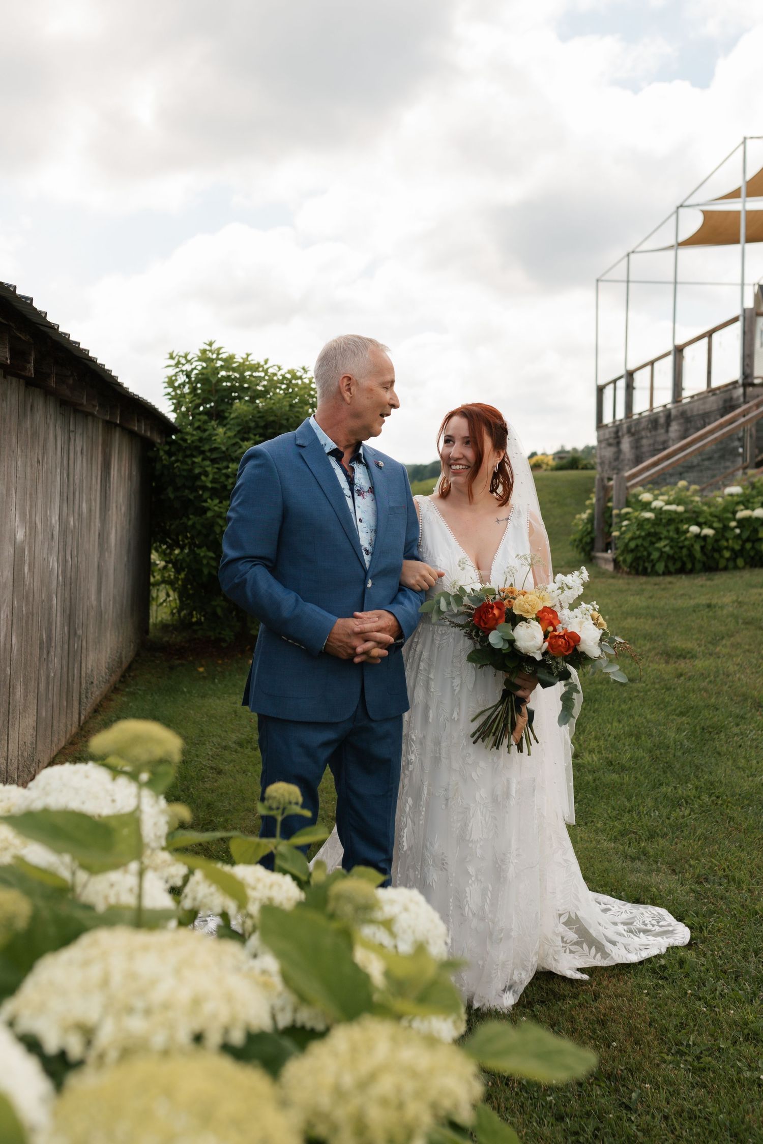 Bride in lace wedding gown holding colorful bouquet walks with man in blue suit at outdoor wedding venue.
