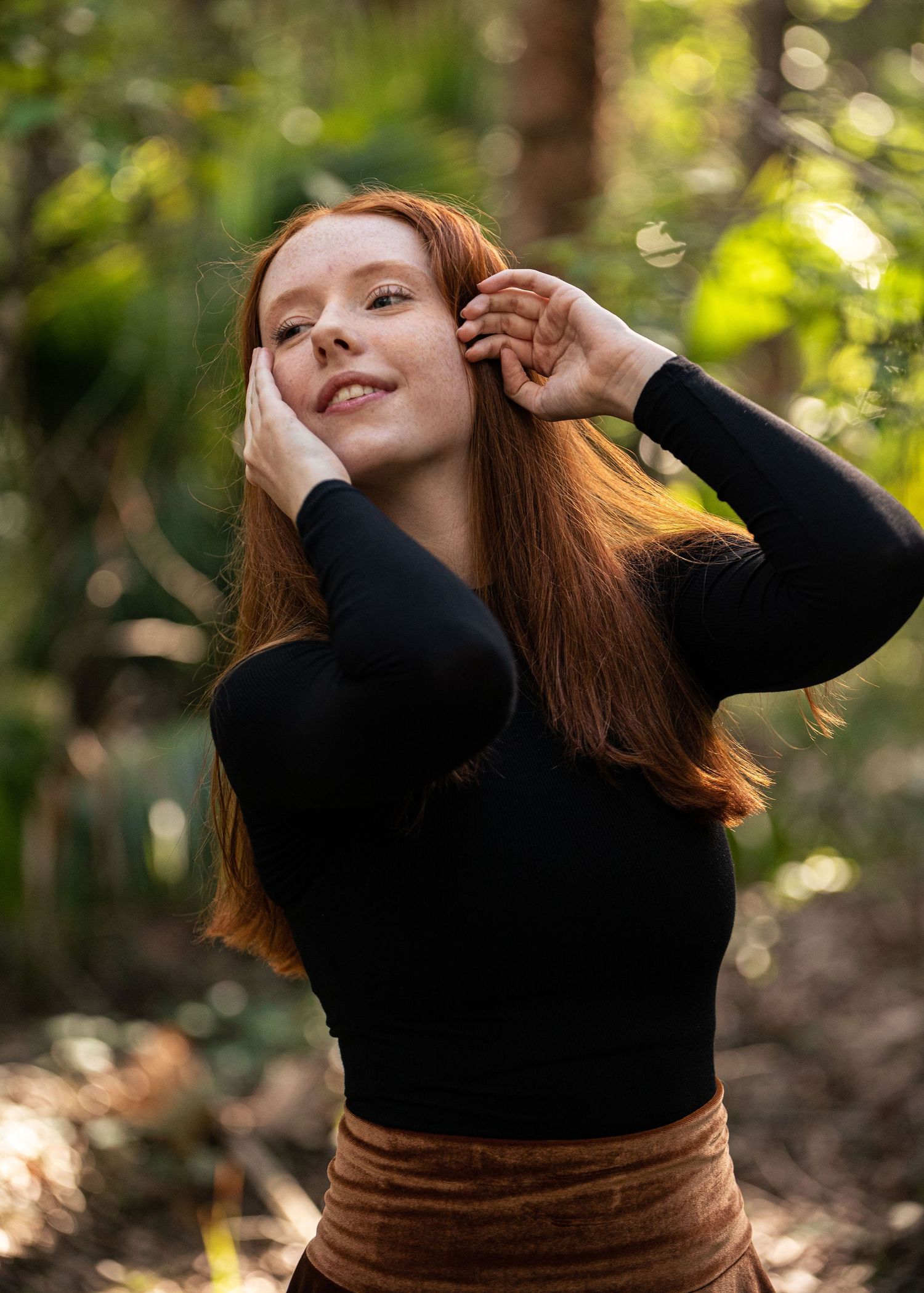 Person in black sweater poses playfully in a sunlit forest setting.
