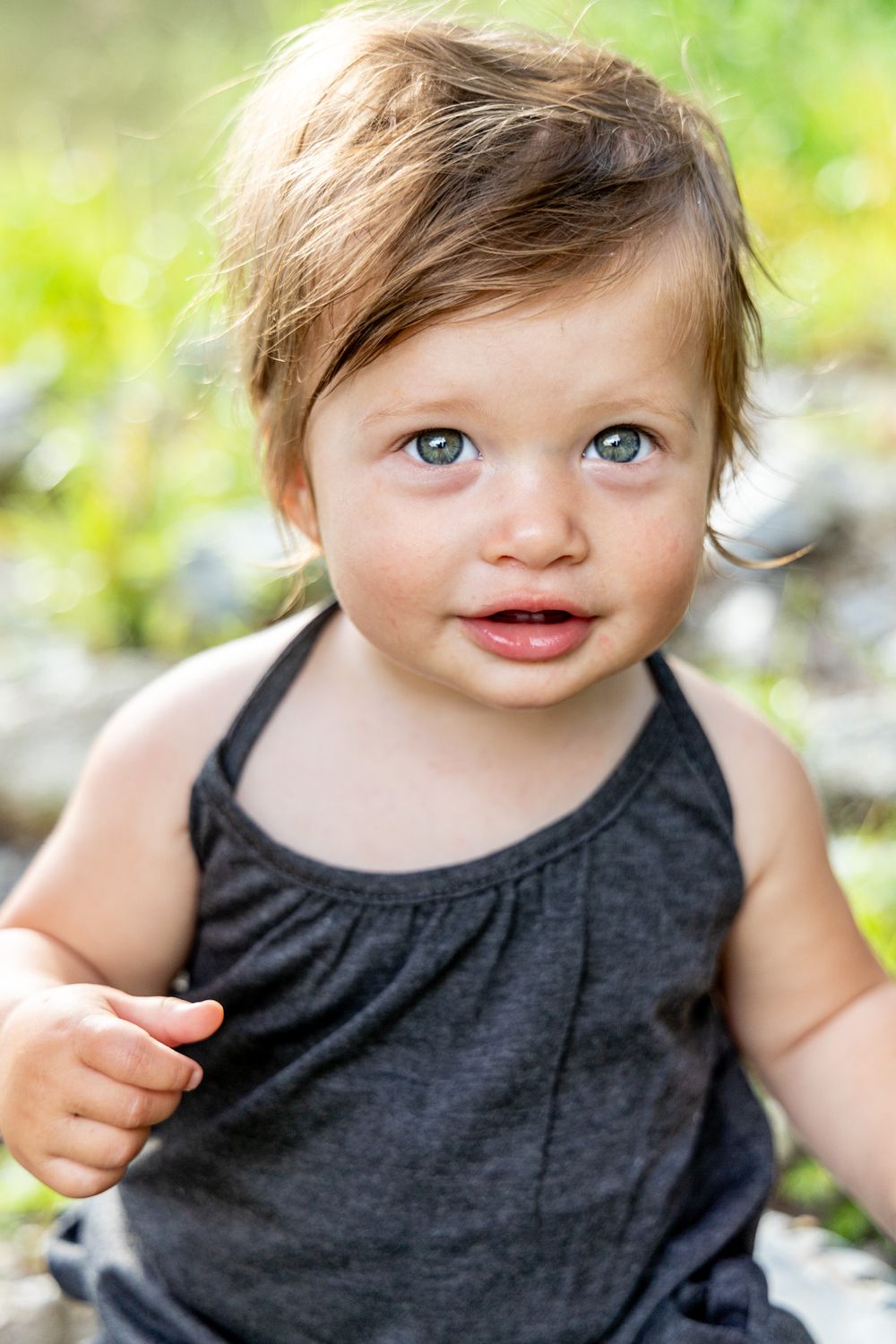 A toddler in black tank top smiles brightly in outdoor garden setting.