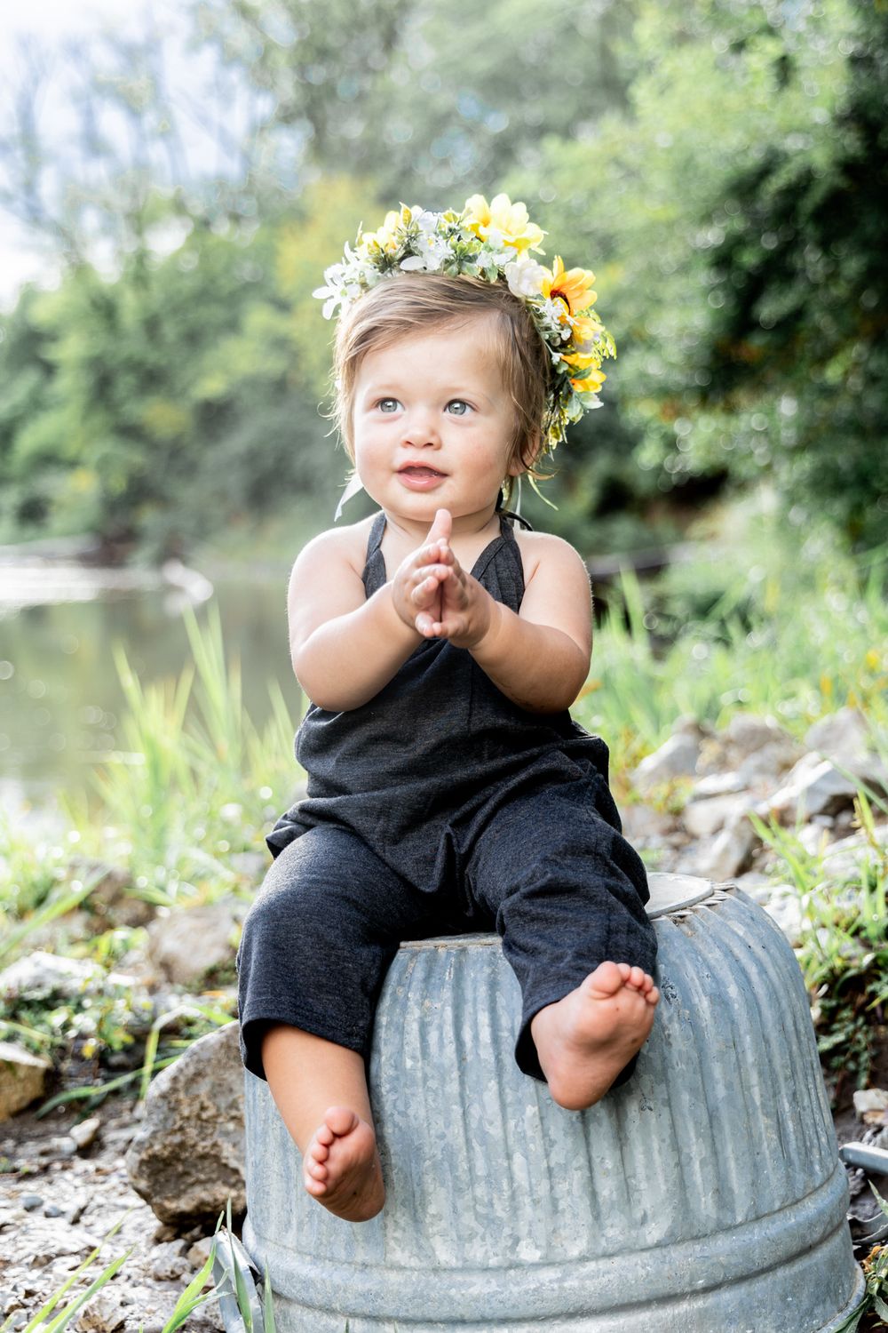 A toddler wearing dark romper and yellow flower crown sits on galvanized container by pond.