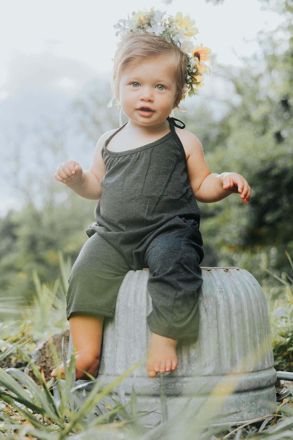 A toddler wearing olive romper sits on galvanized metal container with floral crown in garden setting.