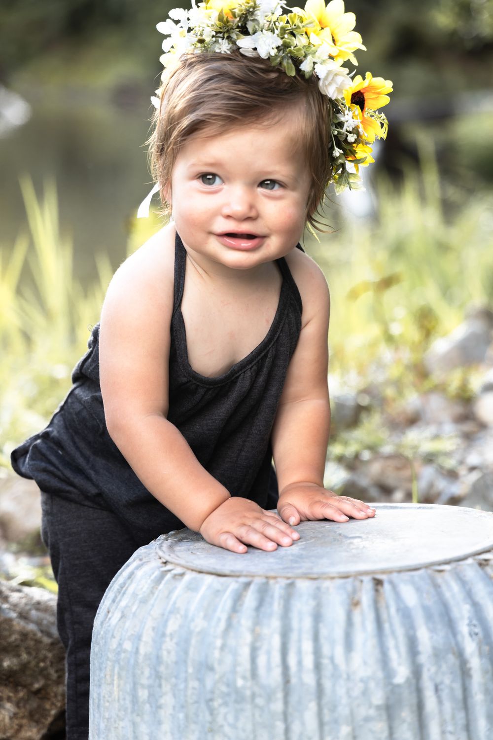 A toddler in black romper with floral crown leans on metal container in nature setting.