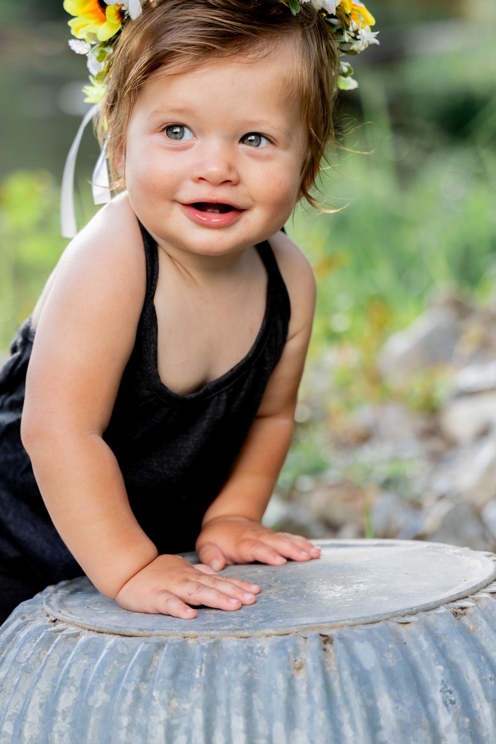 A toddler wearing yellow flower crown and black tank top smiles while leaning on metal surface.