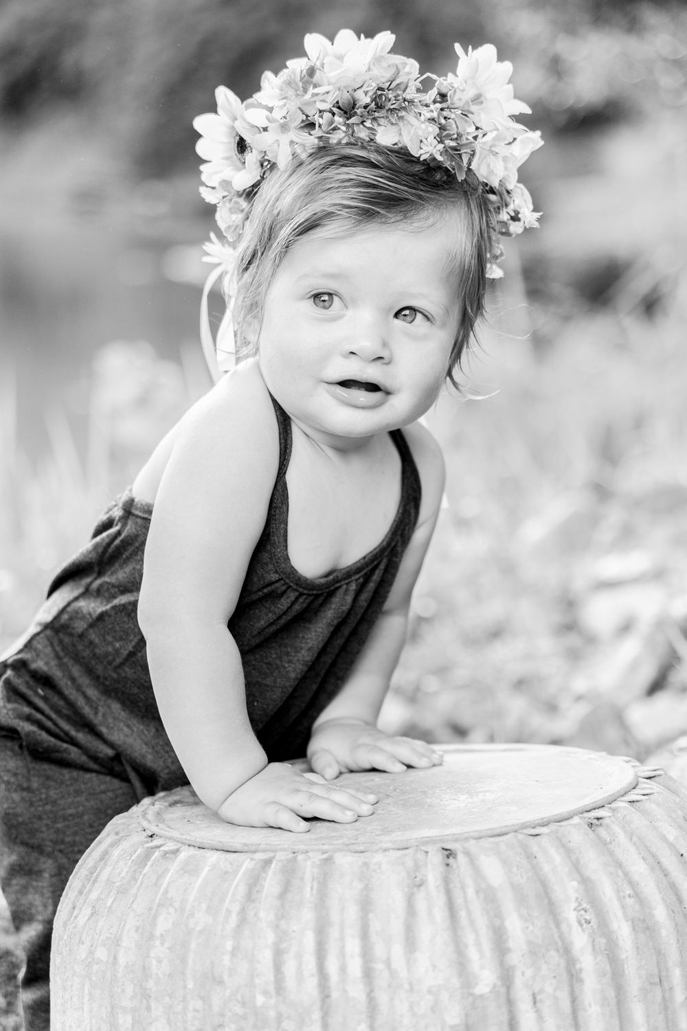 Black and white portrait of a small child wearing a flower crown and dark outfit leaning on a white pumpkin.