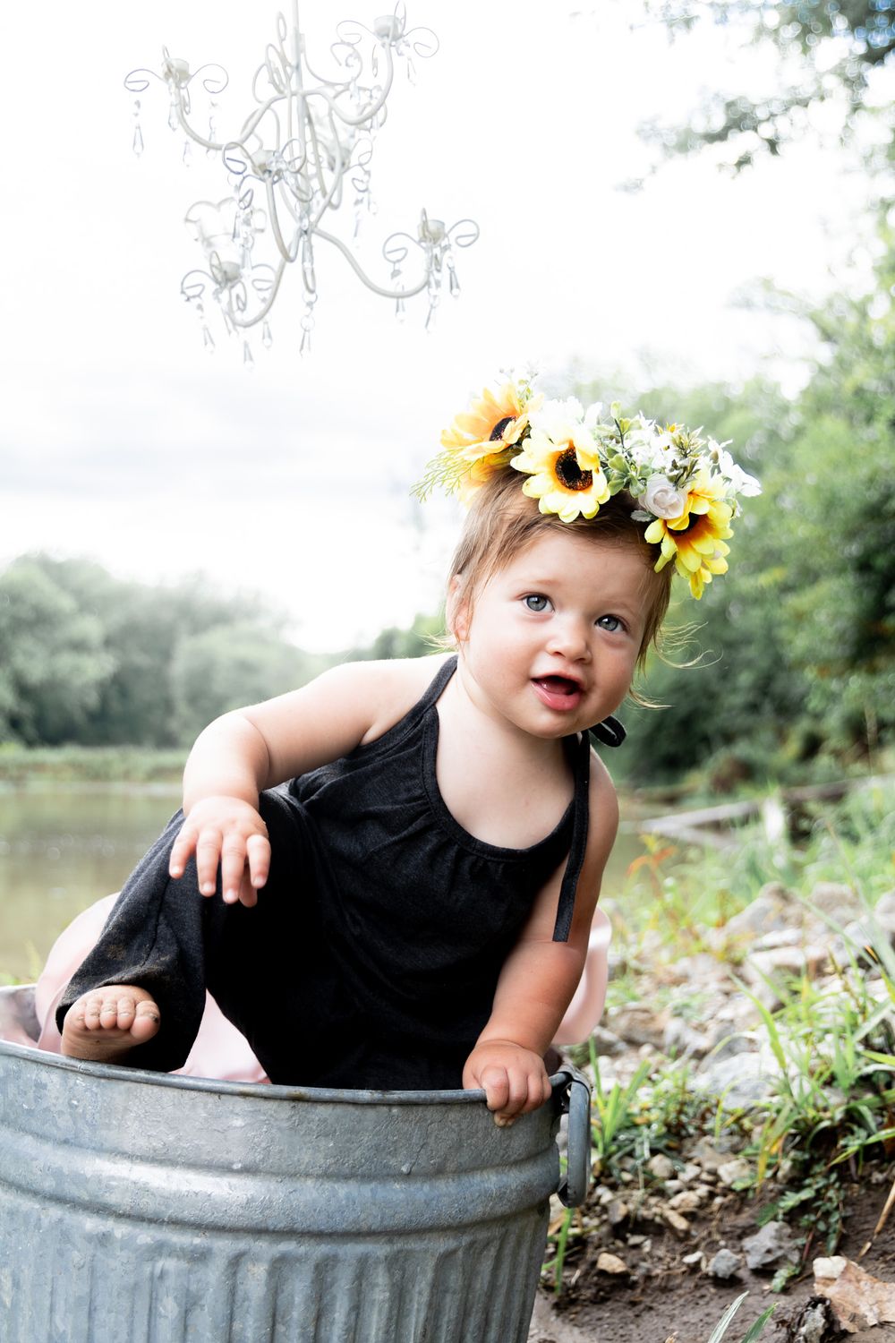 Small child in black outfit with sunflower crown playing in metal bucket by pond.