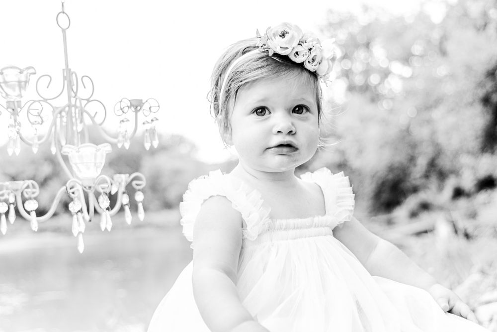 Black and white photo of small child in white dress under ornate chandelier.
