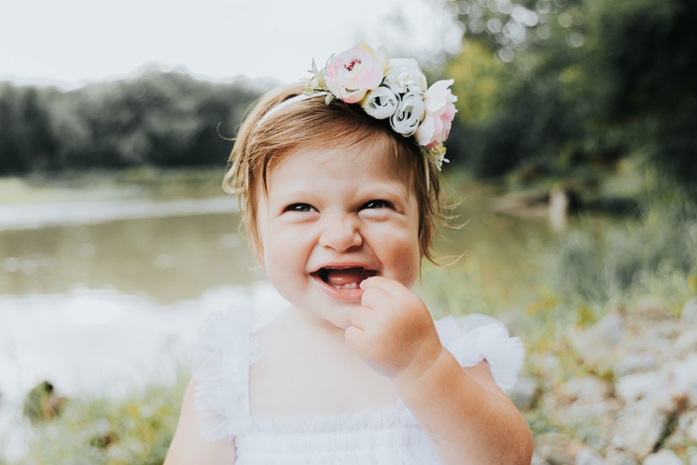 Close up outdoor portrait of small child in white dress wearing floral headband by pond.