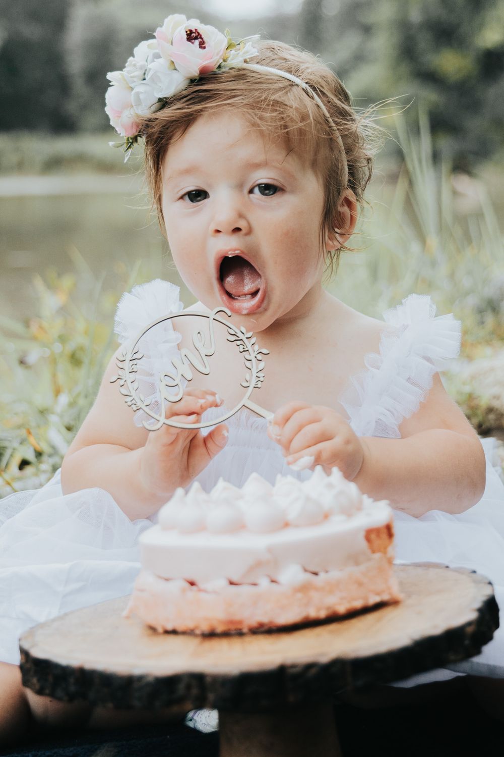Small child in white dress with floral crown excitedly looking at pink cake outdoors.