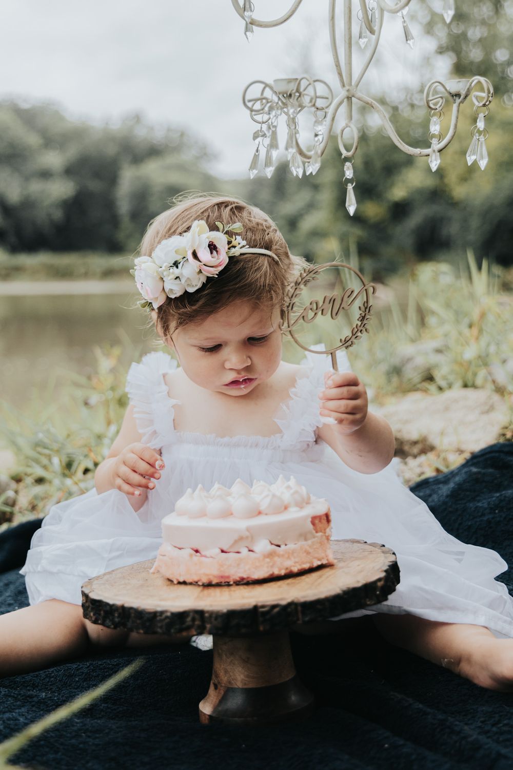 Small child in white dress examining cake decoration while sitting with pink cake outdoors.