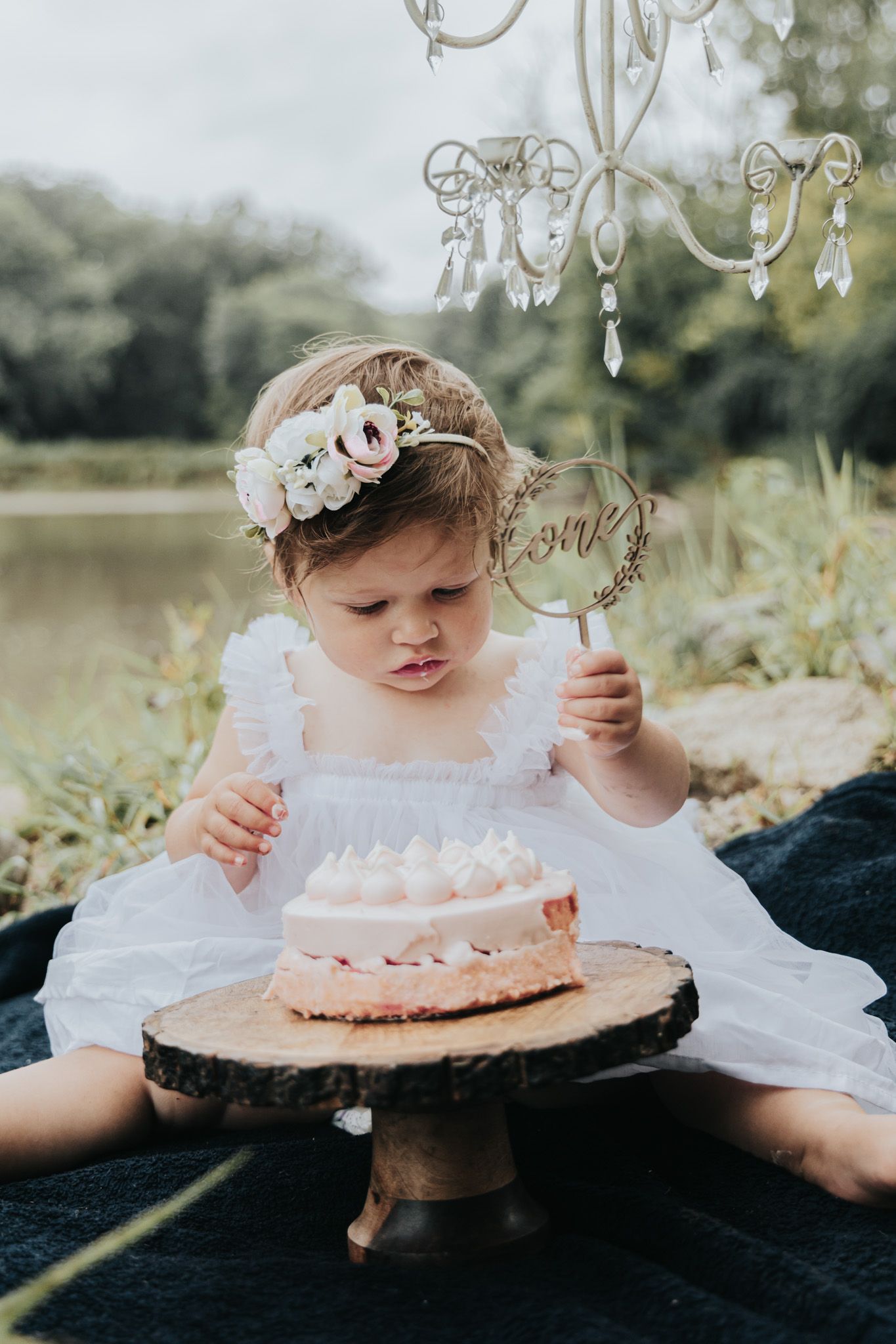 Small child in white dress examining cake decoration while sitting with pink cake outdoors.
