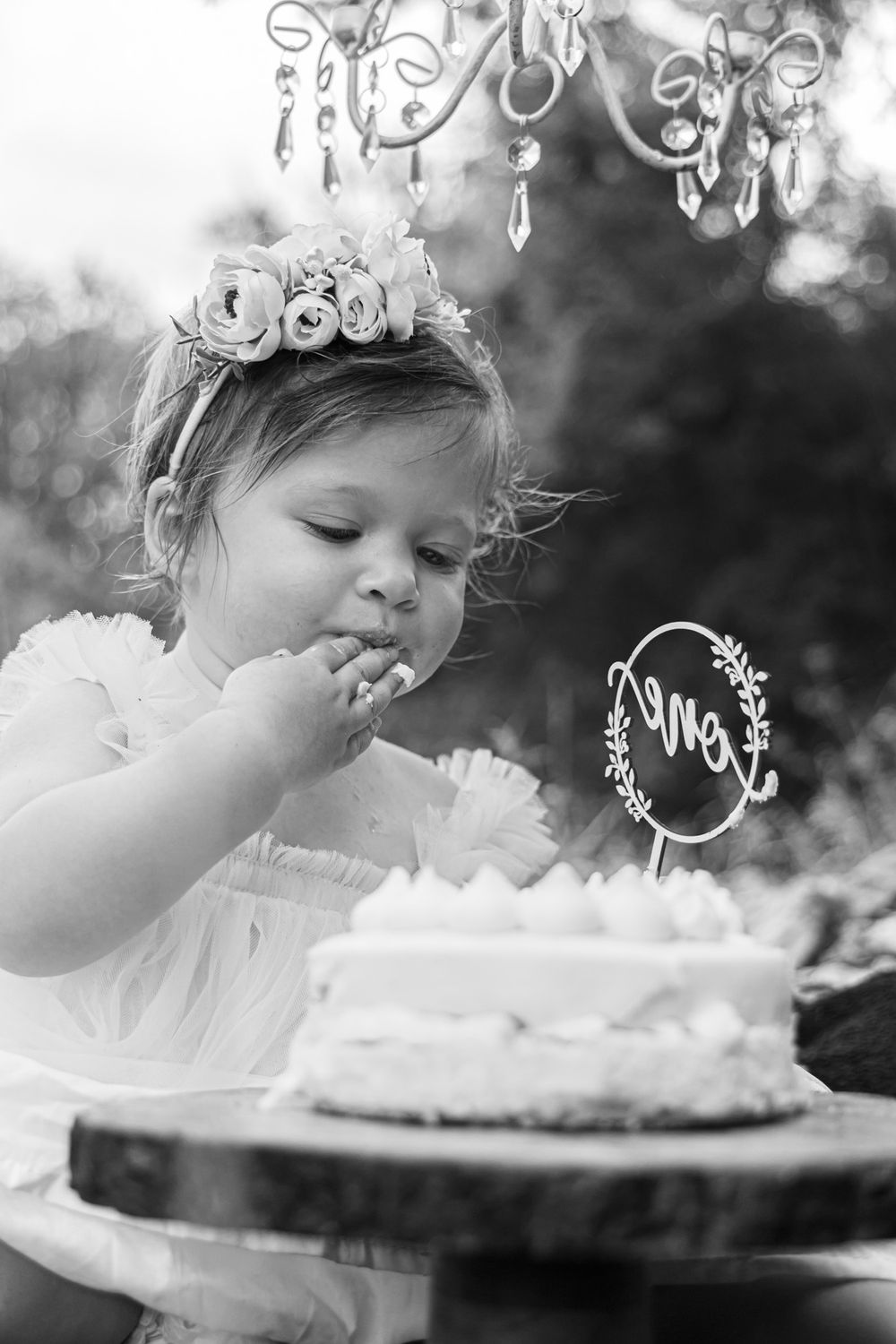 Black and white image of small child in white dress tasting cake under hanging chandelier.