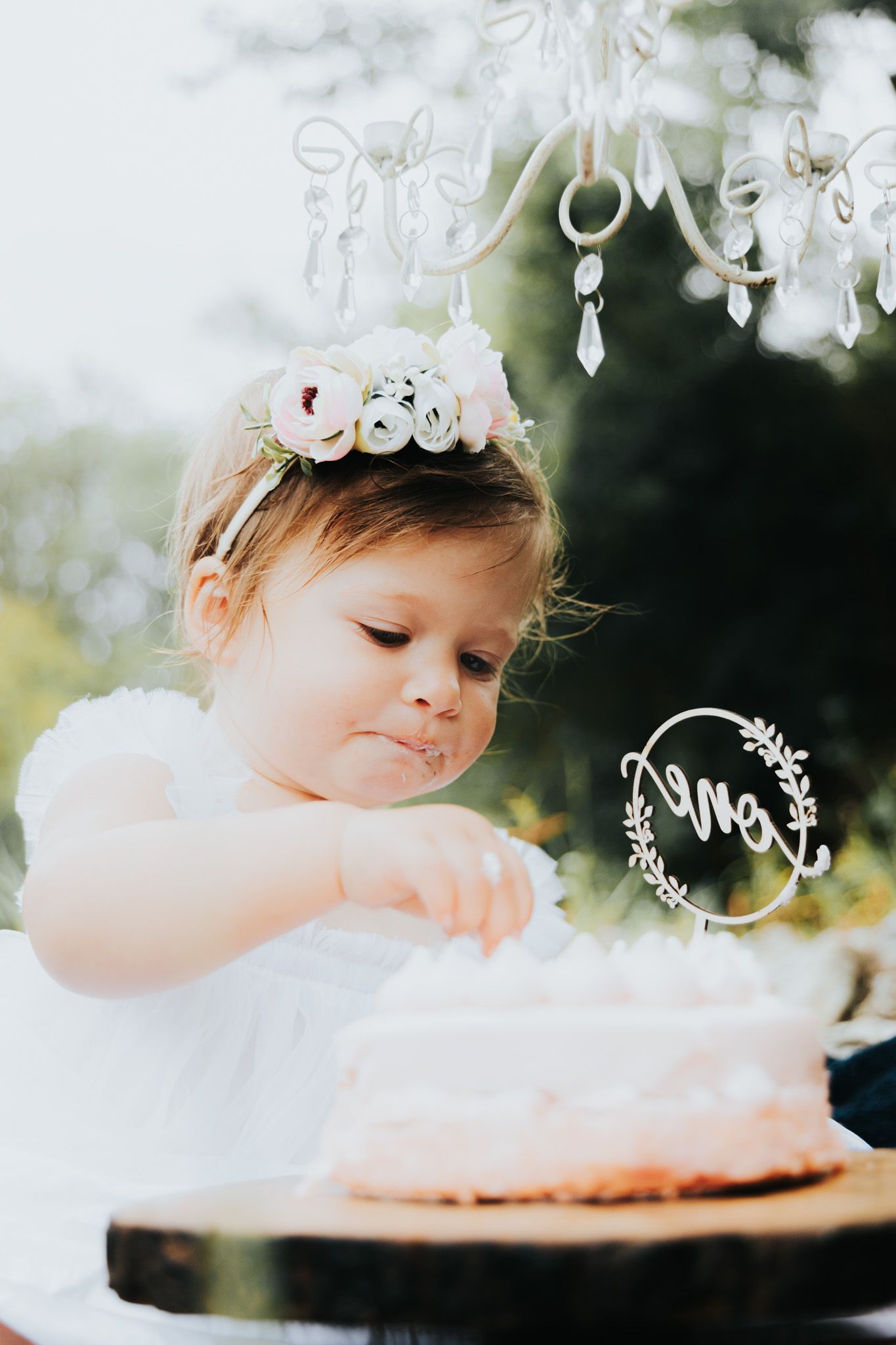 Small child in white dress reaching for pink cake under decorative hanging lights outdoors.