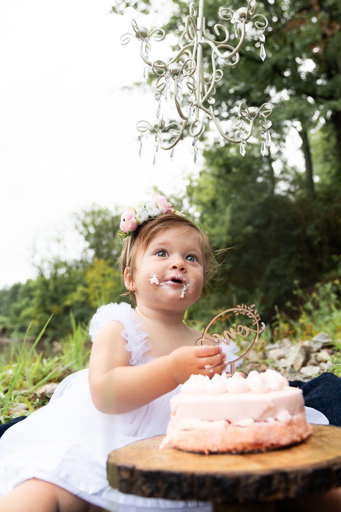 Small child in white dress smiling at cake with decorative topper in outdoor garden setting.