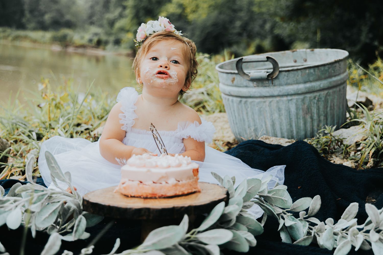A white dress wearing figure sits outdoors with a pink cake and rustic galvanized bucket in a garden setting.