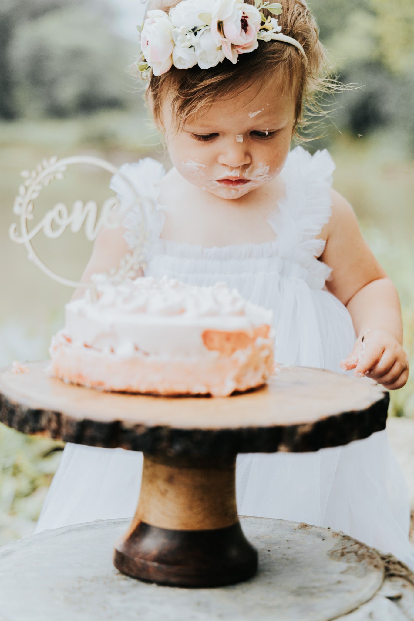 A delicate floral crown adorns a white dress as small hands reach for a pink cake on wooden stand outdoors.
