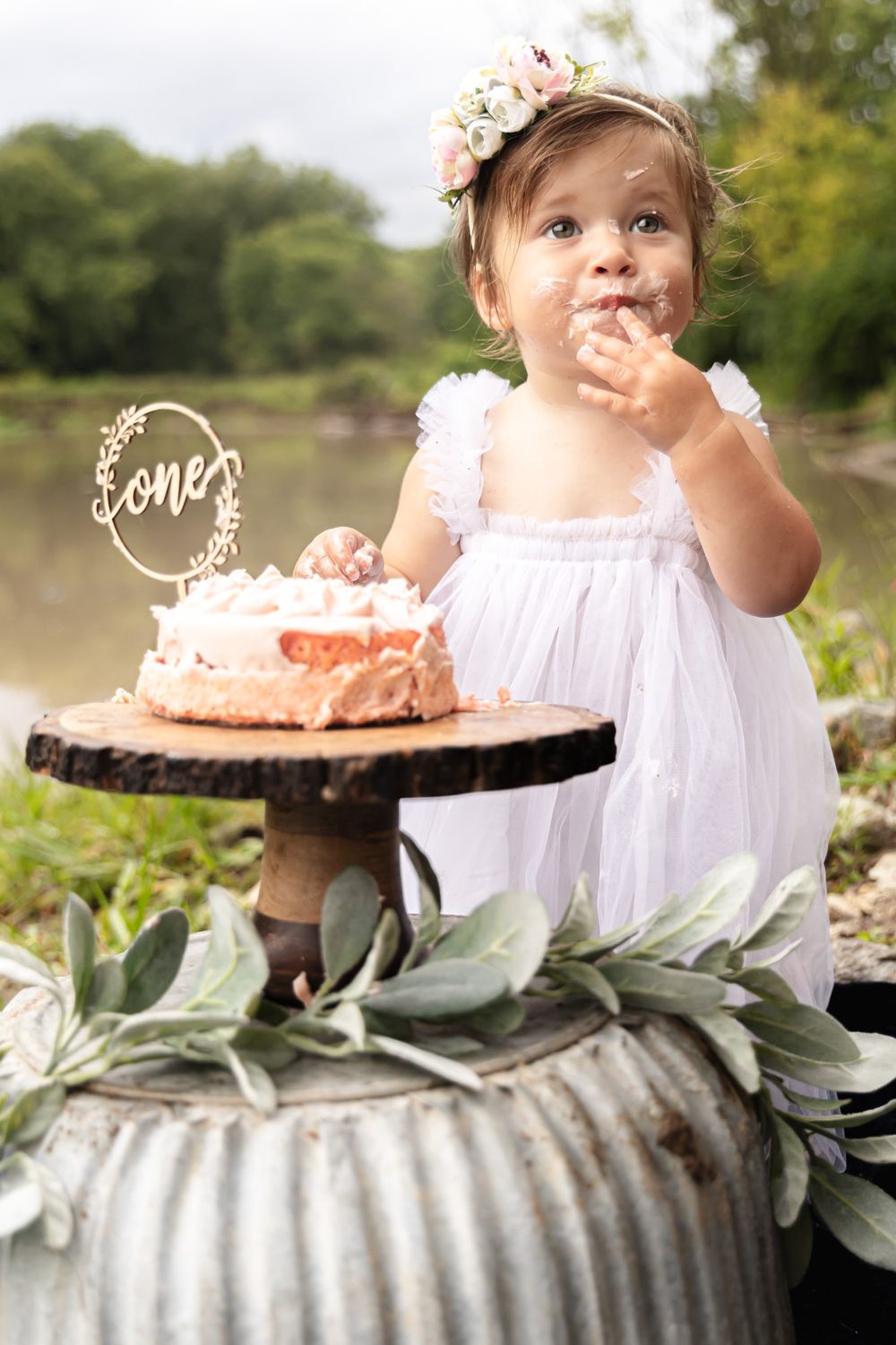 A toddler in white dress tastes birthday cake on rustic metal stand with floral decorations.