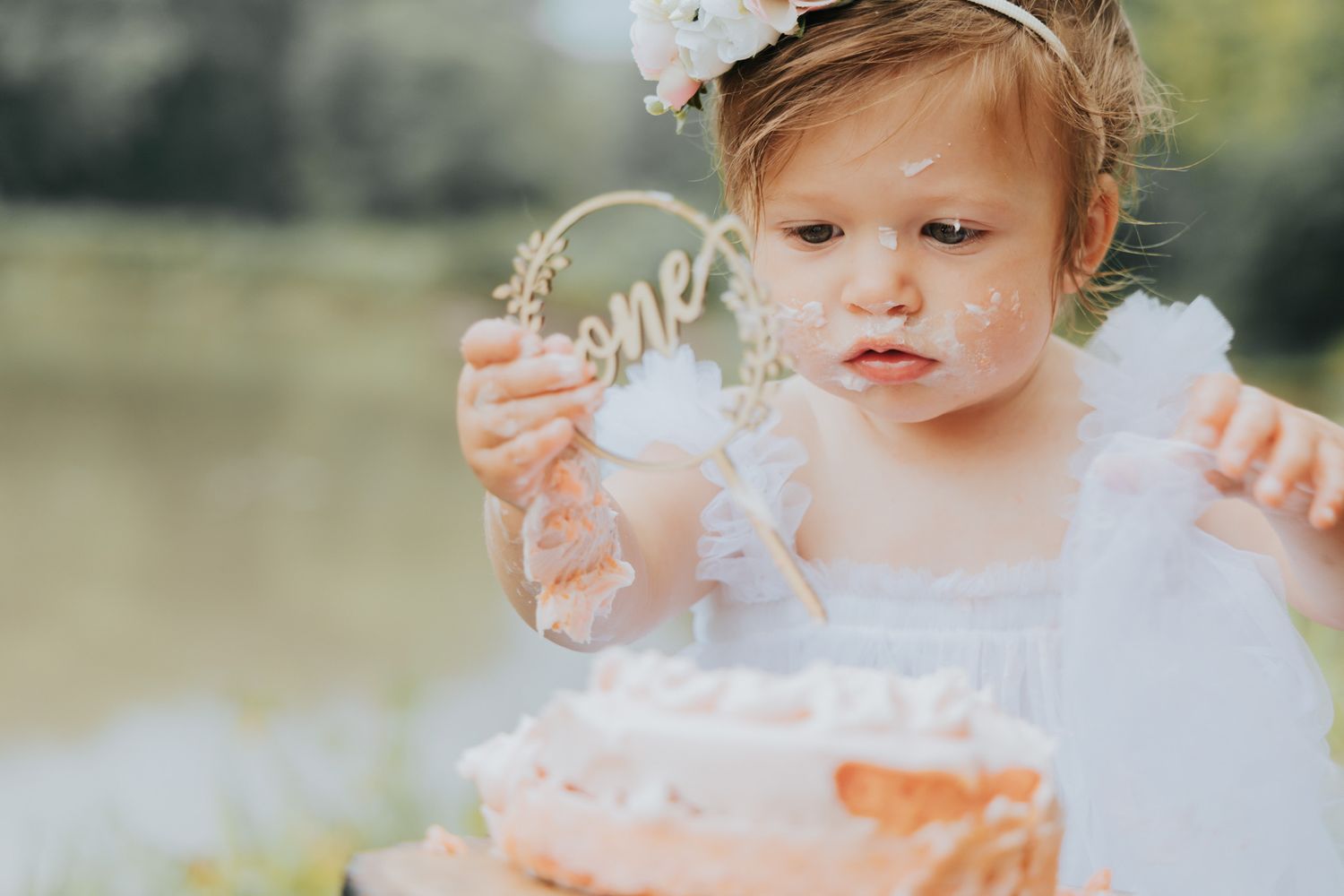 Small hands reach towards a pink frosted cake in an outdoor garden setting.