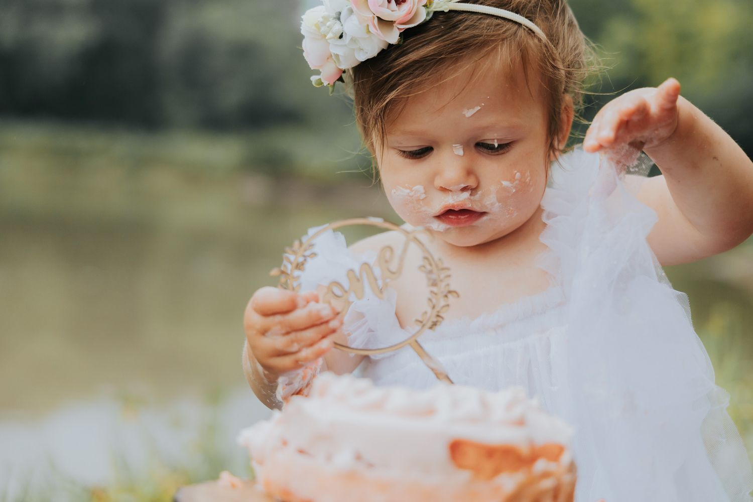 Tiny fingers explore a pink frosted cake while wearing a white ruffled dress outdoors.