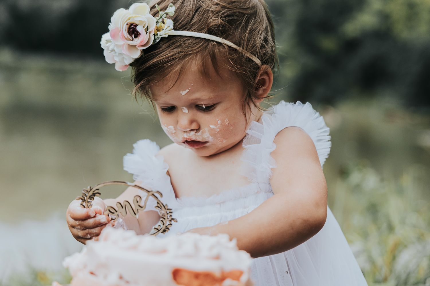 Small hands touch a pink cake while wearing a white dress and floral headpiece in nature.