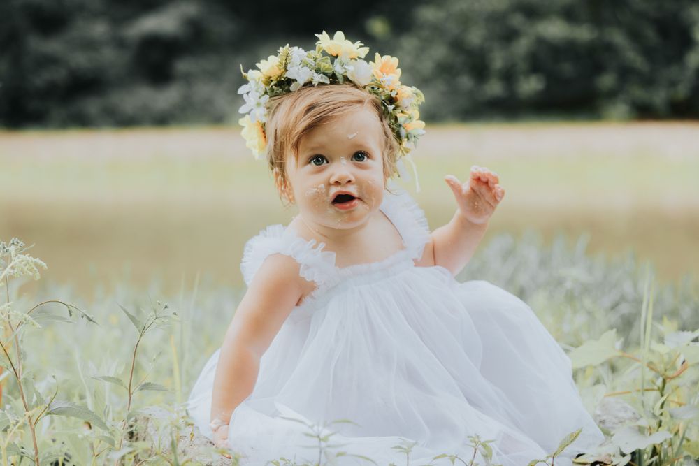 A toddler in white dress plays in wildflower field wearing flower crown.