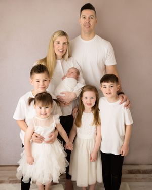 A family wearing coordinated white and cream outfits poses together against a neutral background for a portrait session.