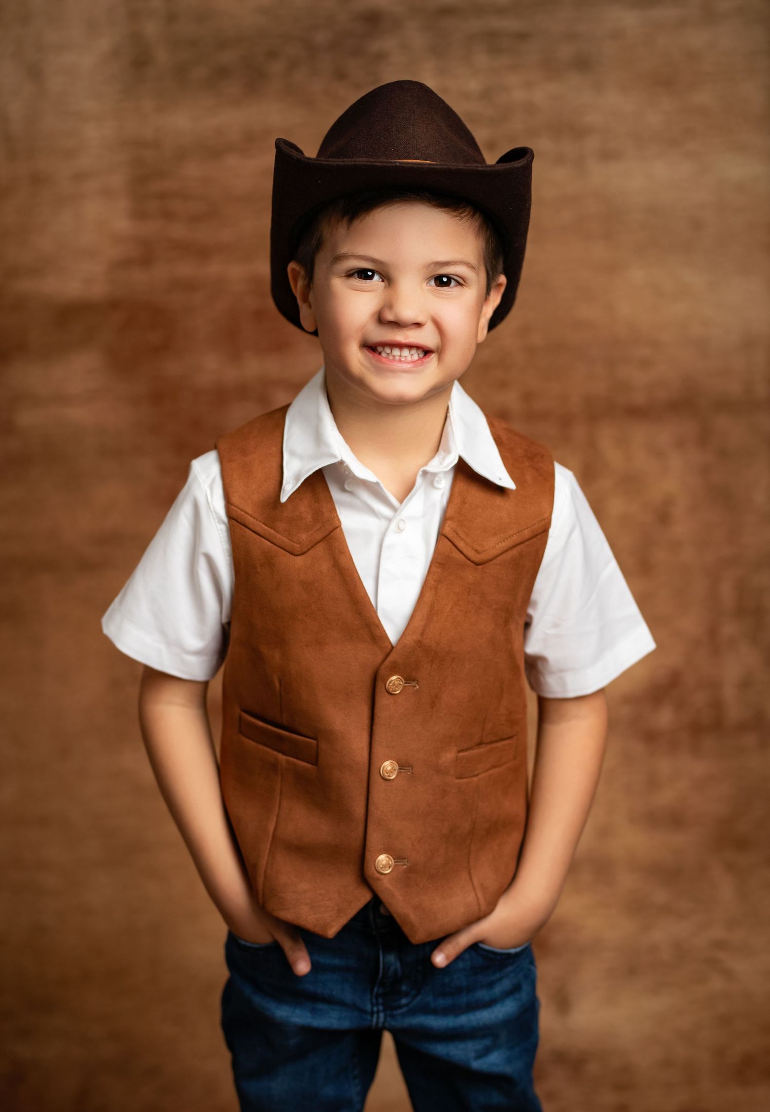 Young child in western cowboy attire with brown vest, white shirt, cowboy hat and jeans against brown backdrop.