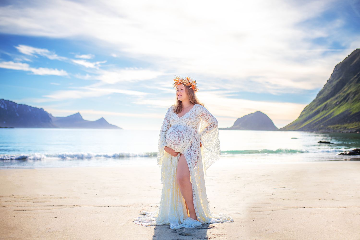 Pregnant woman in white lace gown with floral crown stands on sunny beach with mountains.