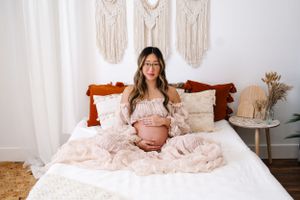 Expecting mother in a soft, ivory maternity gown, sitting is a boho studio holding her belly during a photoshoot.