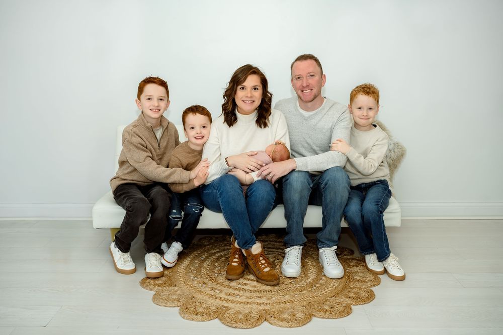 A family sits together on a round jute rug against a light gray wall in a bright studio setting.