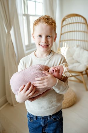 A series of four photos showing a child in a white sweater and jeans holding and smiling at a newborn baby.