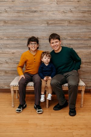 Three children sitting together on a wooden bench against rustic wood paneled wall.