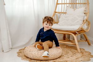 A child sits on a round woven pouf in a cozy corner with a hanging rattan chair and jute rug.