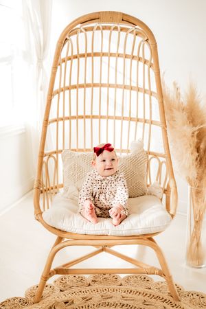 Baby wearing polka dot outfit sits in rattan peacock chair with white cushions and pampas grass decor.