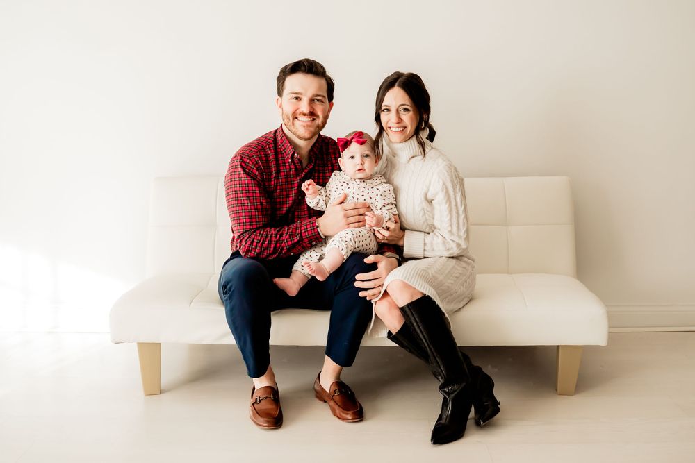 A couple sits on a white couch with a baby wearing a red dress in a bright minimalist studio setting.