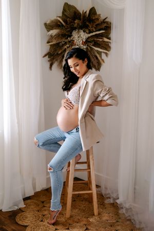 Expecting mother in a soft, ivory maternity gown, sitting in a boho studio holding her belly during a photoshoot.