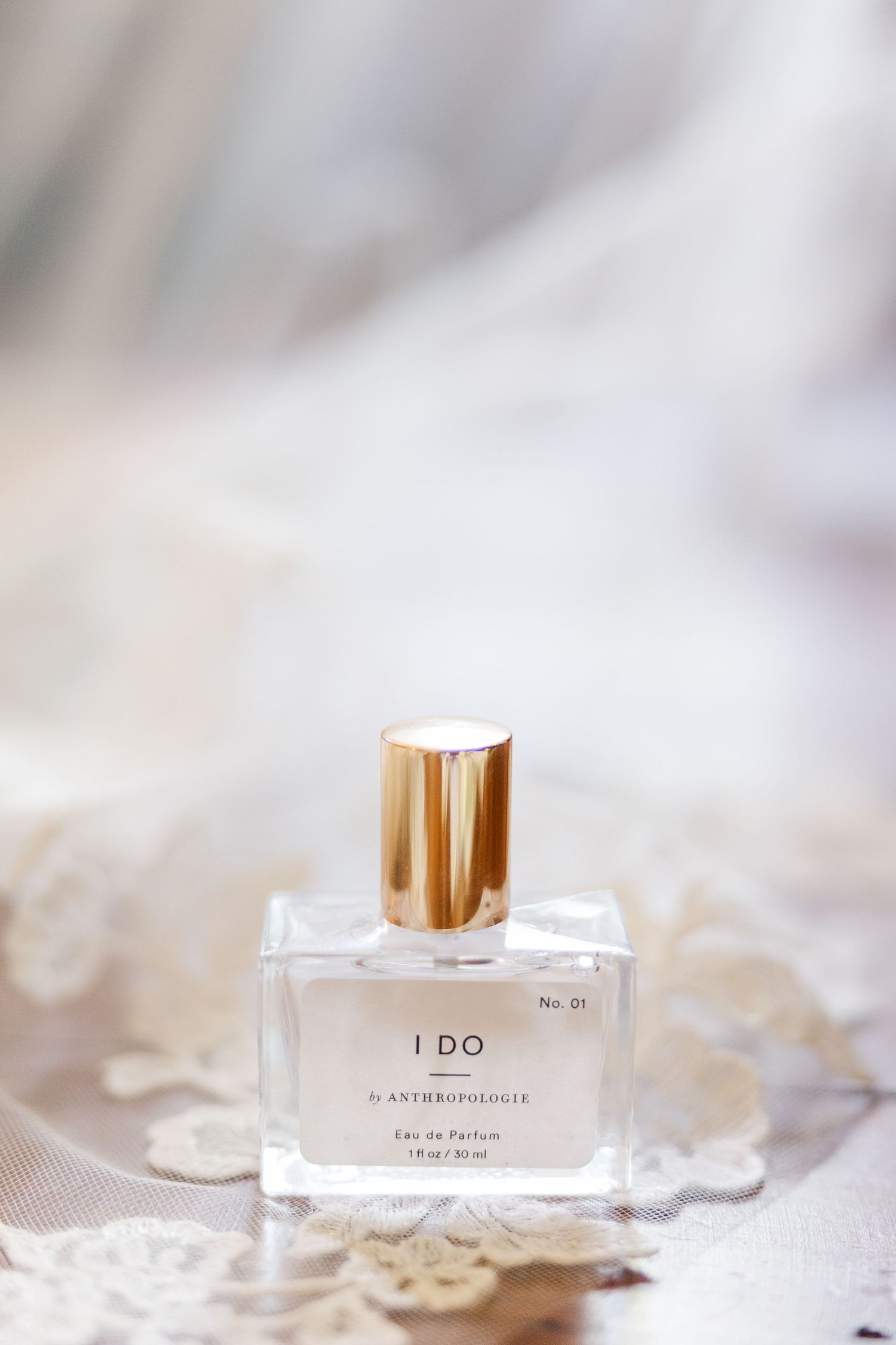 A bottle of 'I Do' perfume sits elegantly on delicate white lace with a gold cap in soft lighting.