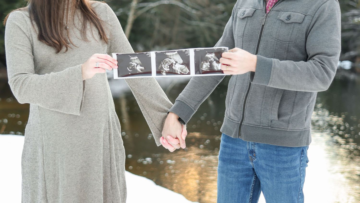 Pregnant couple holding ultrasound photos together outdoors in snowy winter setting by a river.