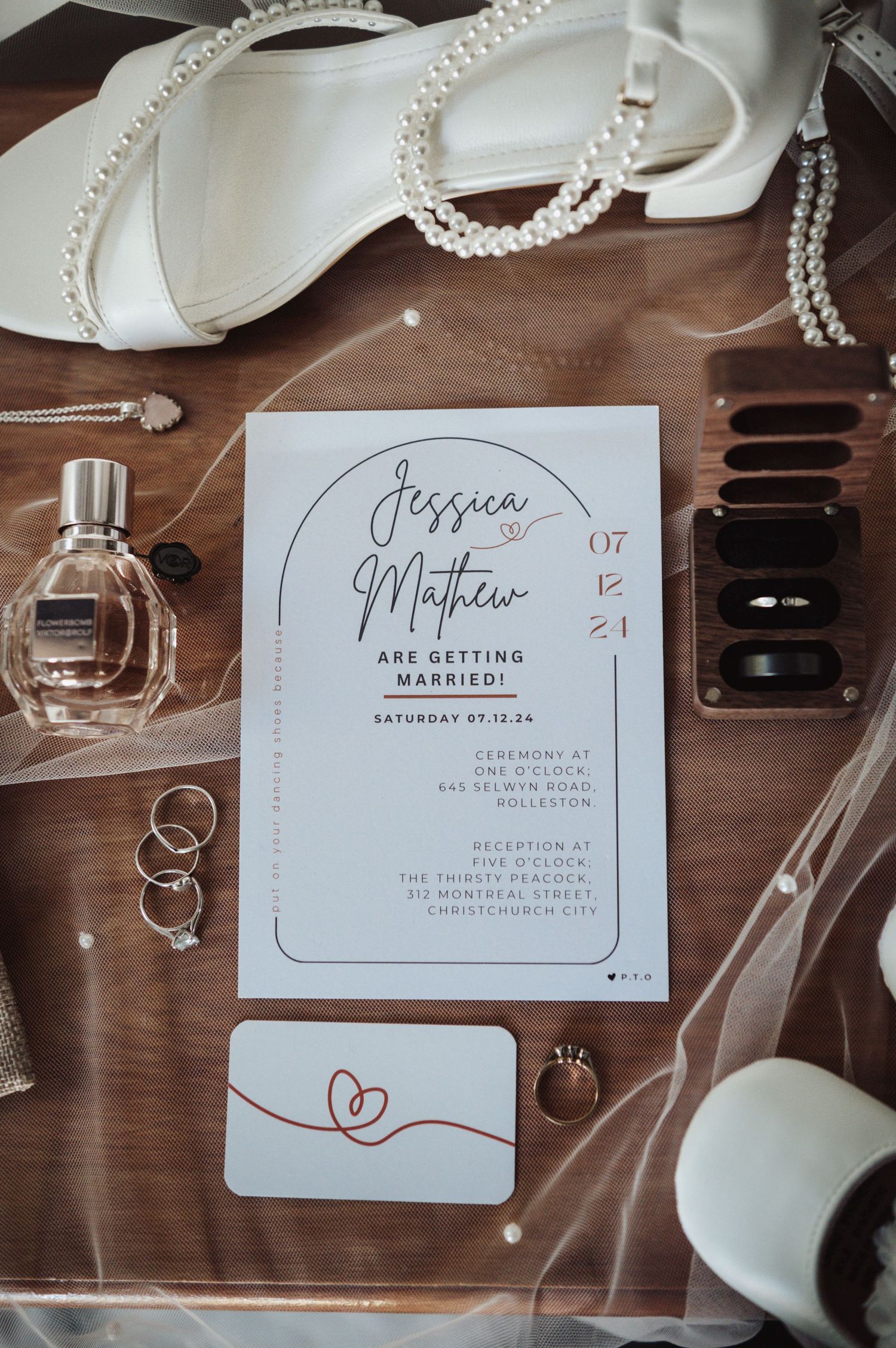 Wedding invitation flat lay with pearls, perfume, and bridal accessories on wooden surface.