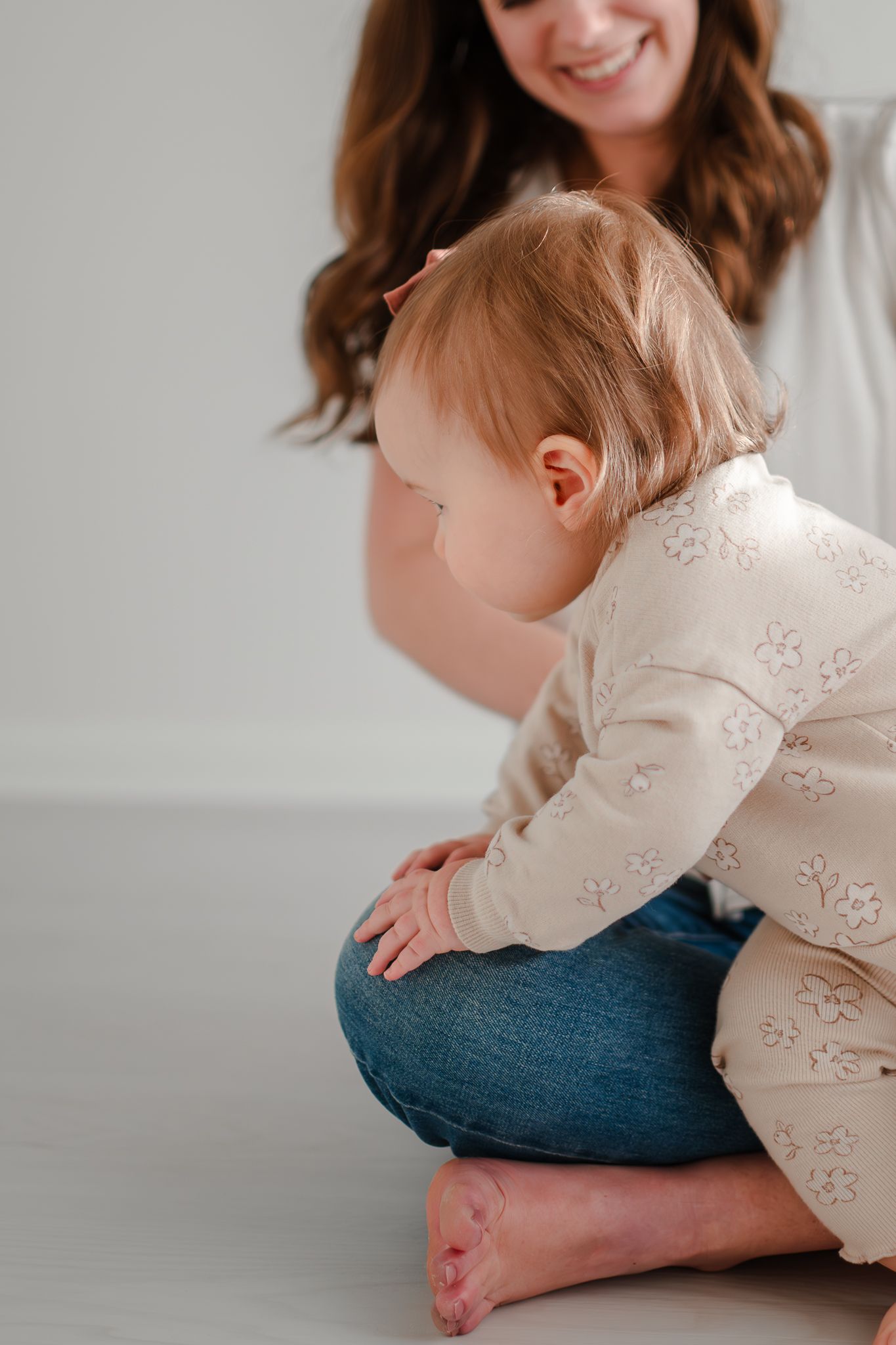 Baby in beige floral onesie crawls on white floor while playing with parent in casual denim.