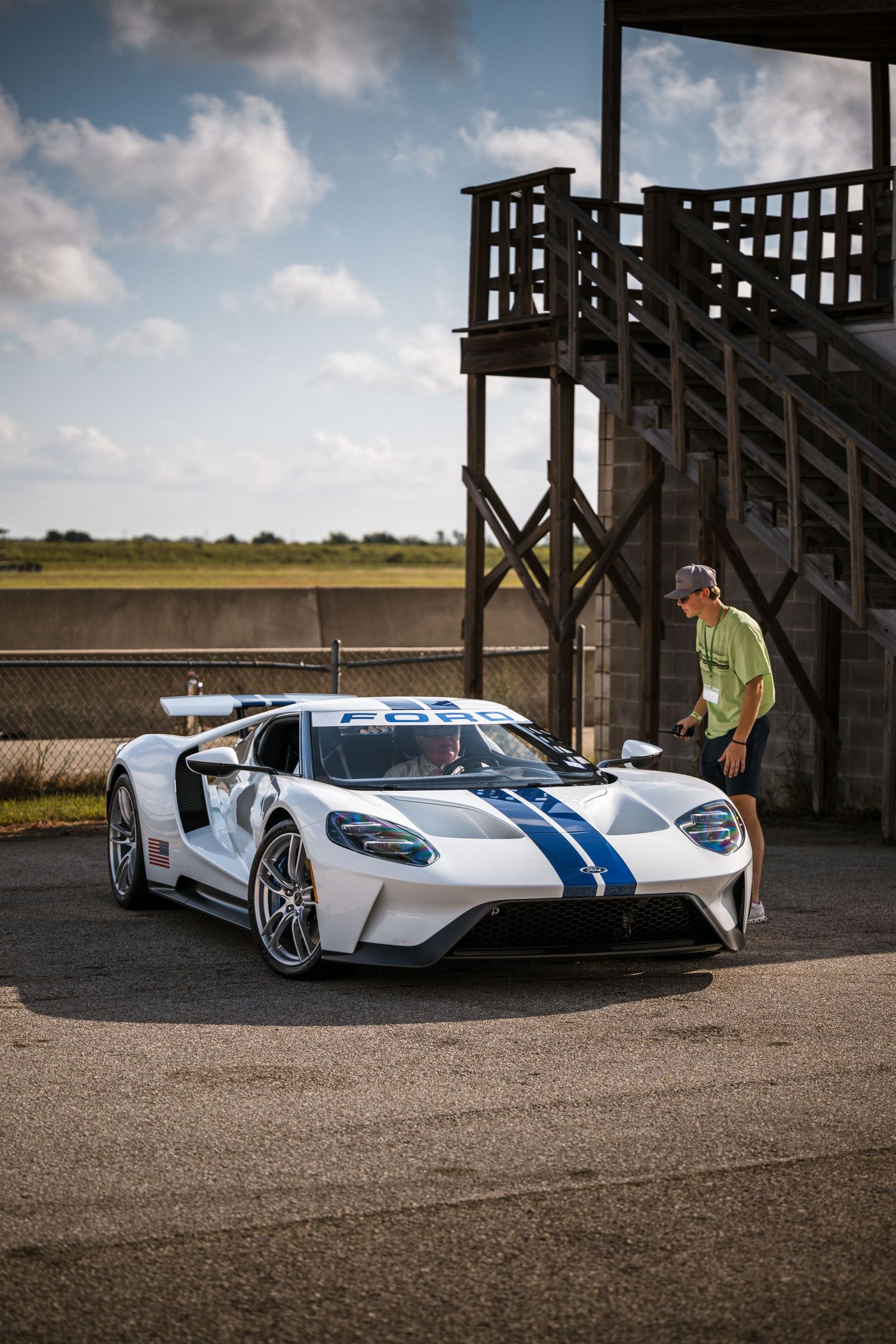 White Ford GT with blue racing stripes parked near wooden structures in rural setting.