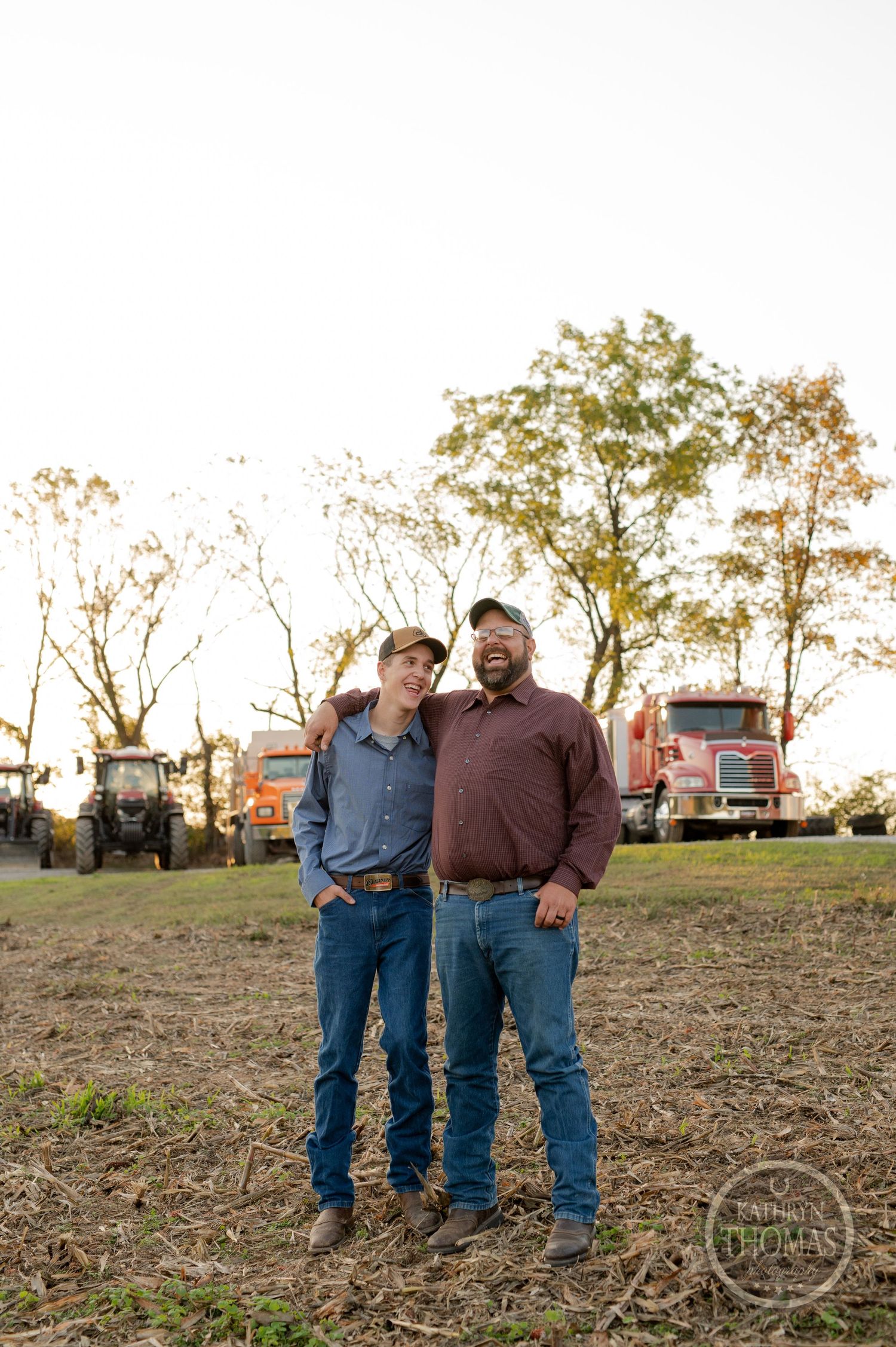 Two people stand together on a dirt field at sunset with farming equipment in the background.