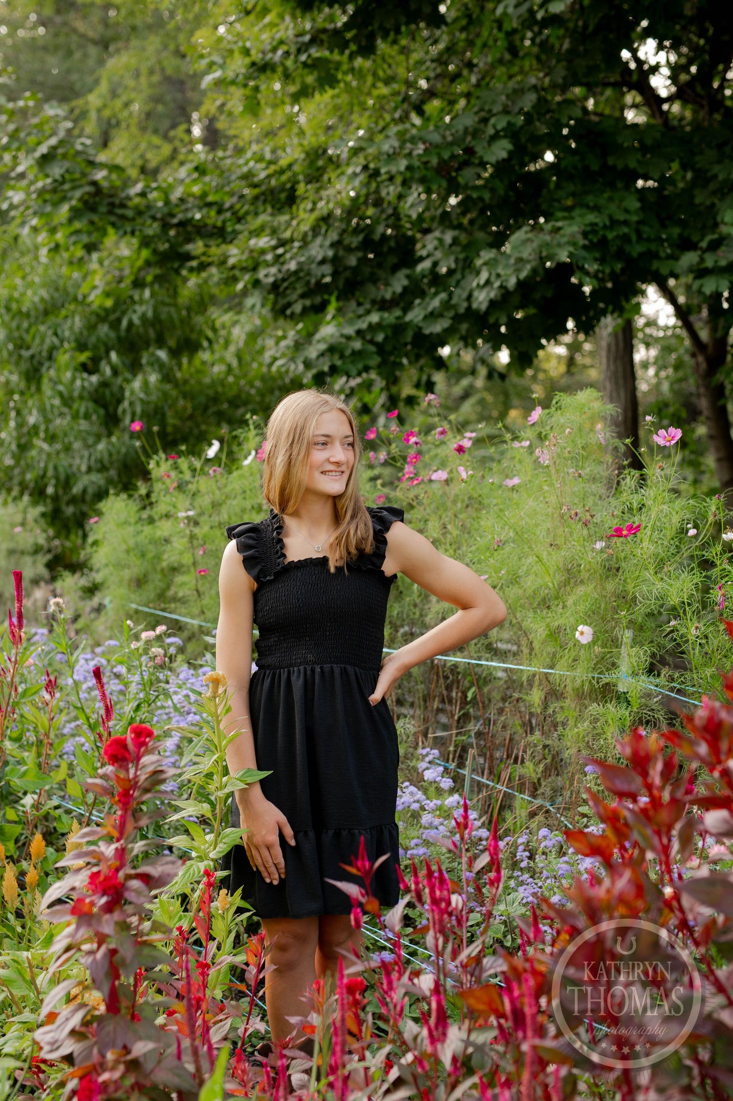 A person poses in a black dress among red cardinal flowers and greenery in a summer garden.